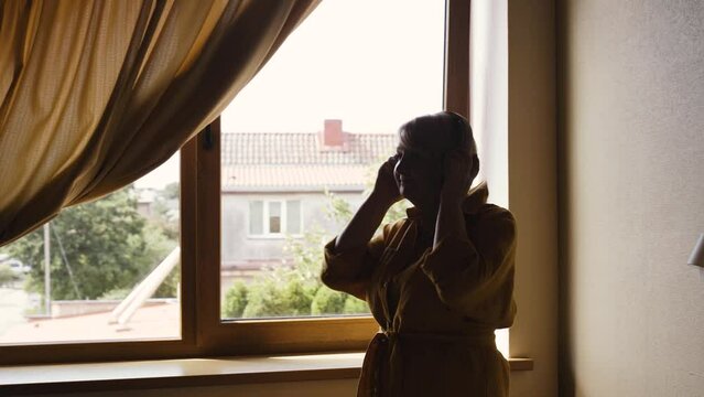 An elderly positive woman stands near the window in the room, listens to music in wireless headphones and dances.