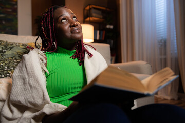 Inquisitive smart afro american woman teacher tutor sitting in the living room at cozy home beautiful young lady holding reading book resting relaxing after hard day