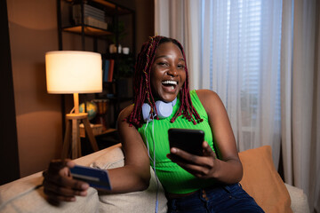 Cheerful dlighted dark skinned black woman wearing in casual bright colorful clothes holding cellphone in hands and new credit cards doing online purchase shopping at weekends - obrazy, fototapety, plakaty