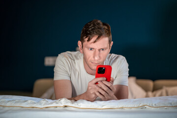 Man uses smartphone lying on bed. Middle aged male severe, unsmiling, uses mobile phone, makes...