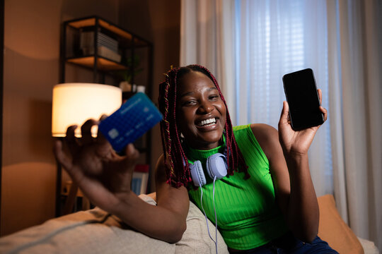 Happy delighted smiley afro american dark skinned woman with colorful hair wearing purple headphones looking at camera holding credit cards showing at camera