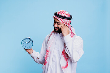 Man wearing traditional muslim clothes holding retro alarm clock and looking at time. Arab person...
