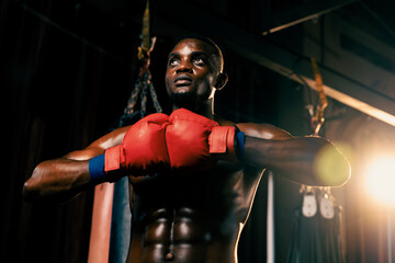Fototapeta na wymiar Boxing fighter posing, African American Black boxer put his hand or fist wearing glove together in front in aggressive stance and ready to fight at gym with kicking bag and boxing equipment. Impetus