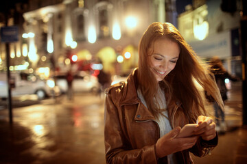 Fototapeta Young Caucasian woman using a smartphone on the sidewalk in London UK at night obraz