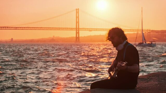 Hipster street musician in black playing electric guitar in the street on sunset on embankment with 25th of April bridge and yacht boat in background. Lisbon, Portugal