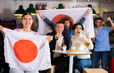 Company of emotive young adult friends rooting for favorite team and waving flag of Japan while...