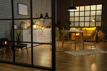 Interior of modern kitchen with glowing lamps, dining table, yellow sofa and white counters at...