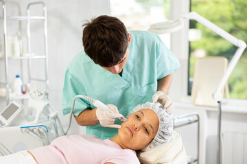 Young cosmetic doctor confidently utilizes hydrogen oxygen cleaning attachment on machine,...
