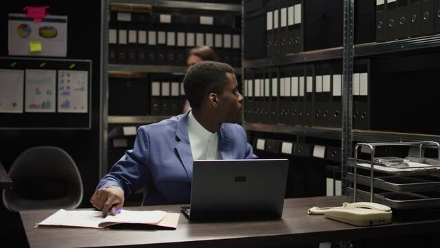 Dedicated male private investigator examines classified documents from shelves. African American policeman cross-checking evidence and statements on wireless computer and case forensic records.