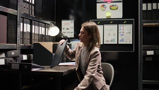 Caucasian female inspector examines confidential case files and records. Young private detective walks into office space filled with clues and evidence, positioning laptop on table and starts working.