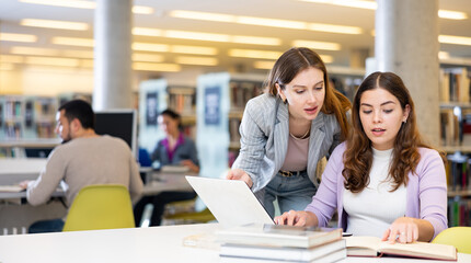 Obraz premium Two young women finding information at library together, working with laptop and books