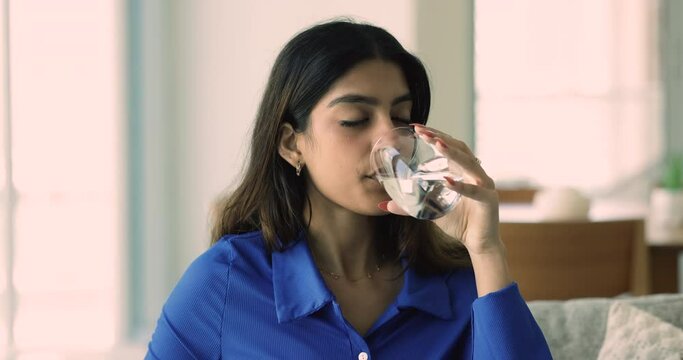 Close up portrait of young Indian woman hold glass with fresh water, looking at camera enjoy natural aqua seated on sofa, makes sip, drink enough liquid per day, take care of health. Healthy lifestyle