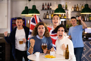 Company of joyful sports supporters waving flag of Great Britain and celebrating victory of...