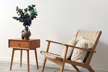 Vase with green eucalyptus branches on end table and comfortable armchair near white wall - obrazy, fototapety, plakaty