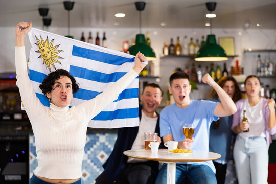 Screaming young adult sports fans rooting for favorite team and waving flag of Uruguay while watching match together in pub. High quality photo