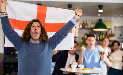 Enthusiastic company of young adult sports fans waving flag of England and supporting national team...