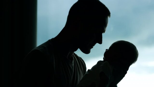 Silhouette of a man holding a little baby. Loving father kissing his adorable child. Window at backdrop.