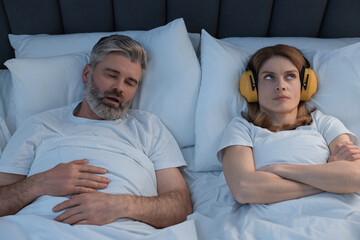 Irritated woman with headphones lying near her snoring husband in bed at home