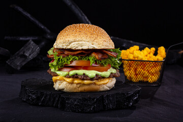 giant hamburger, with a small iron basket containing French fries, on a table with black fabric and wooden items with texture and black color,