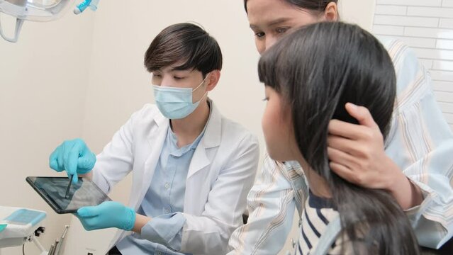 Asian male pediatric dentist explains teeth X-ray scan to girl and her mother for dentistry hygiene from tablet in dental clinic, professional orthodontic oral doctor in a kid healthcare hospital.