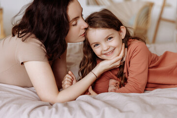 Beautiful young mother kisses her happy cute little daughter lying on the bed in the bedroom. Mom and daughter spend time together at home in casual clothes.