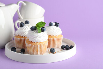 Plate of tasty cupcakes with blueberries on lilac background