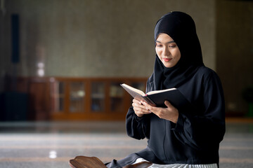 The image of an Asian Muslim woman in the Islamic religion in hijab in black color. Sitting reading the Quran and having a happy smiling face Staying in a beautiful mosque out of respect for God.