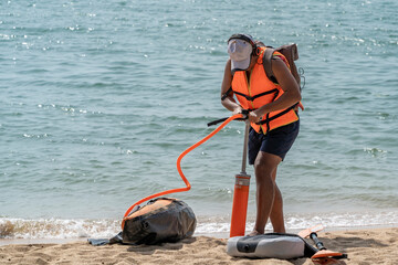 A man in a life jacket, tilting his head, pumps air into a boat that lies on the seashore. A paddle...