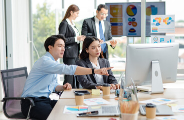 Asian professional successful male businessman mentor supervisor in casual outfit sitting smiling teaching helping female businesswoman intern colleague in formal suit working via computer in office.