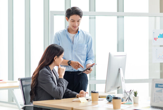 Asian professional successful male businessman mentor supervisor in casual outfit standing smiling teaching helping female businesswoman intern colleague in formal suit working via computer in office