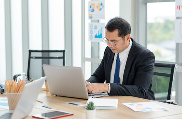 Asian professional successful male businessman manager entrepreneur in formal suit and glasses sitting at working laptop computer at desk in company office