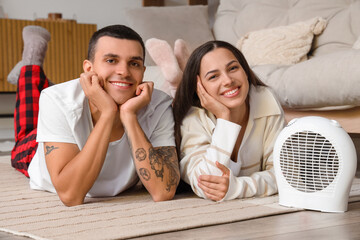 Young couple warming near electric fan heater at home