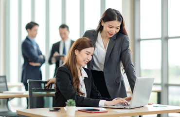 Asian professional successful female businesswoman supervisor mentor in formal suit standing smiling pointing help advising new colleague working with paperwork document at working station in office