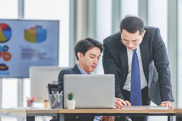 Asian professional successful general manager in formal suit teaching business to male businessman colleague working with laptop notebook computer learning new job in company office meeting room