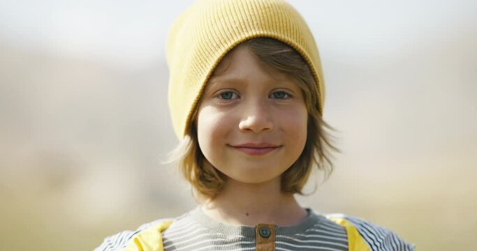 Smiling child 5-6 years old outside. Close-up portrait of child in yellow cap. Blond boy looks into frame. Look thoughtful calm look slight smile, dimples on cheeks. Sunlight. Childhood and serenity