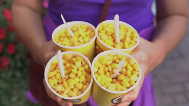 A woman's hand holds four paper cups with boiled corn.