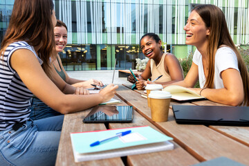 Multiracial university students laughing outdoor in college campus studying and doing homework using laptops.