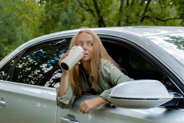 Smiling young woman looking from car window and drinking coffee or tea from reusable thermos cup. Local solo travel on weekends concept. Exited woman explore freedom outdoors in forest. Unity with