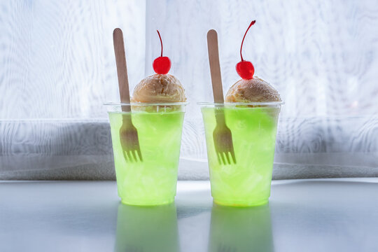 Two cold green apple flavored soda in a clear glass topped with a scoop of vanilla ice cream side the window.