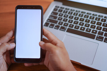 Close up shot of a young asian man in casual clothes using his smartphone and laptop computer at his desk in his home office
