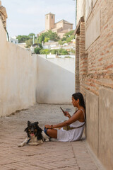 A young woman in a white dress is sitting on the street, checking her mobile phone, while her dog lies comfortably next to her