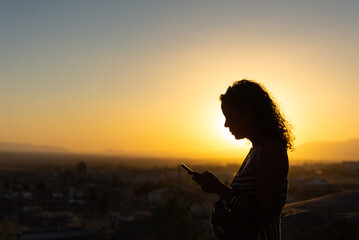 Fototapeta premium Silhouette of a curly-haired woman checking her mobile phone against the backdrop of a cityscape bathed in the warm light of the setting sun