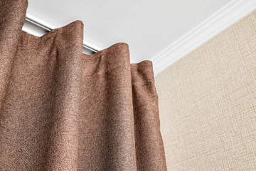 Curtains made of brown matting fabric hang on the ceiling cornice near the wall papered with a matting texture or rough canvas under a white plasterboard ceiling with ceiling baseboards bottom view.