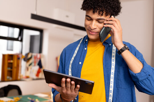 Happy biracial male fashion designer using tablet and smartphone in studio