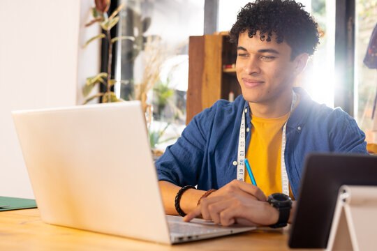 Happy biracial male fashion designer using laptop and tablet at desk in studio