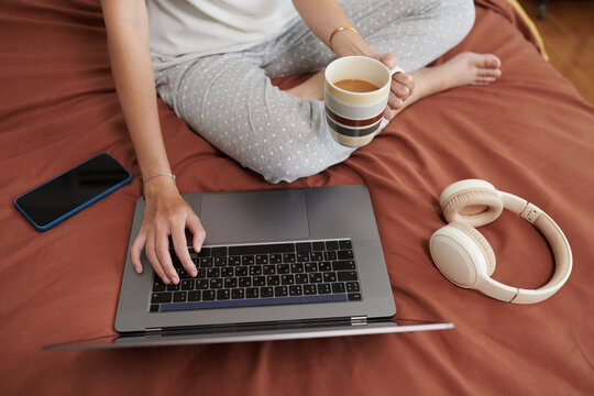 Cropped image of woman in pajamas sitting on bed, drinking coffee and checking e-mails