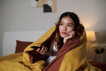 Young woman wrapped in duvet sitting on bed and switching tv channels