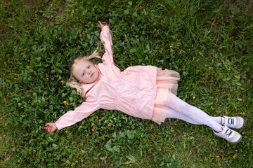 Little blonde girl in pink dress and jacket is lying on green grass with her arms outstretched to the sides. Top view.