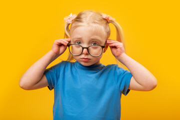 Upset little blond girl adjusts her glasses and looking straight. Portrait of girl in blue T-shirt...