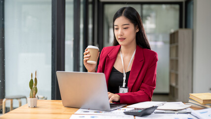 An attractive Asian businesswoman is sipping coffee while working on her laptop at her desk.
